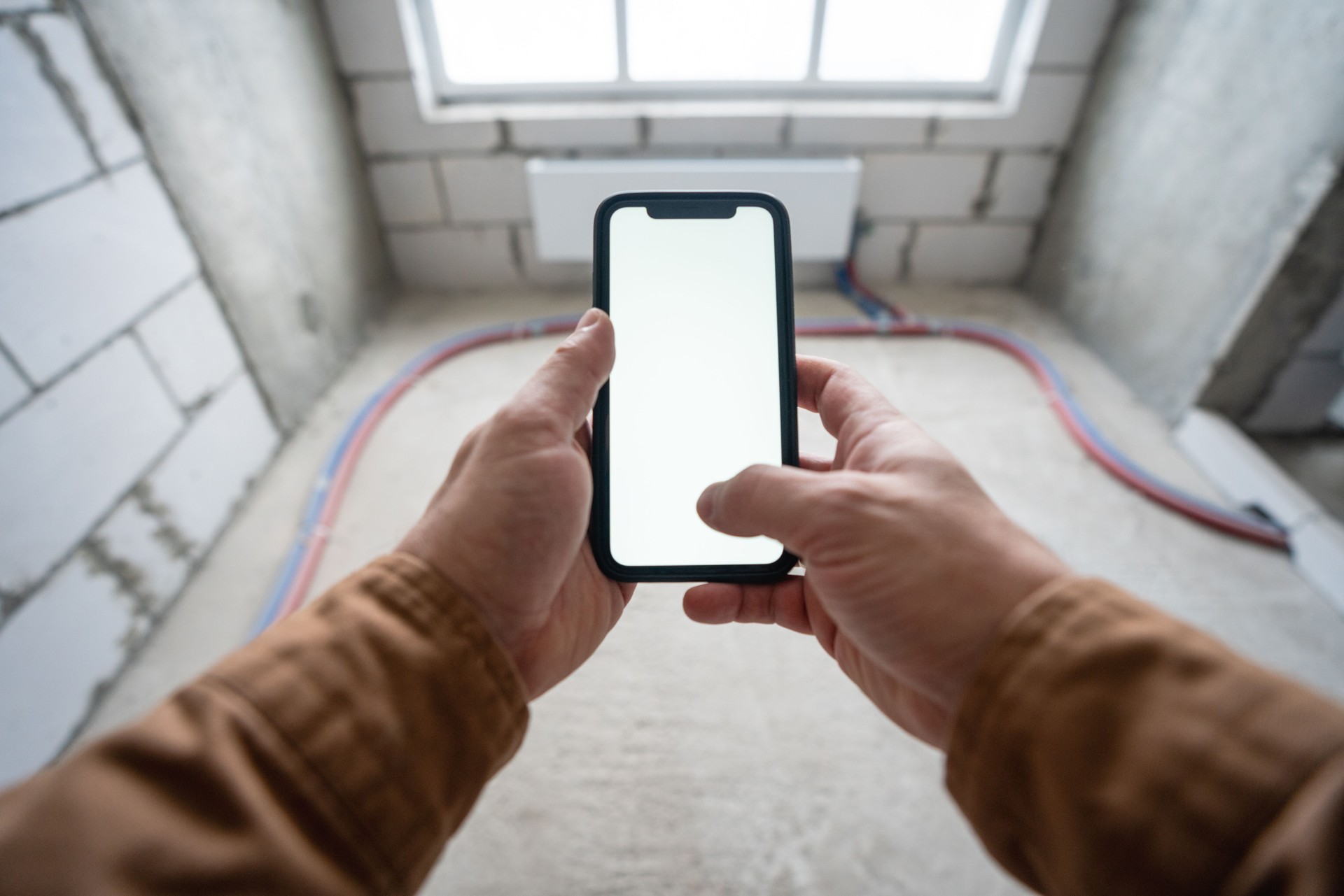 Smartphone with blank white screen in male hands and an empty new apartment without renovation on the background